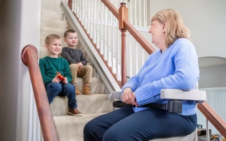 a woman riding an indoor stair lift with kids sitting on the steps watching