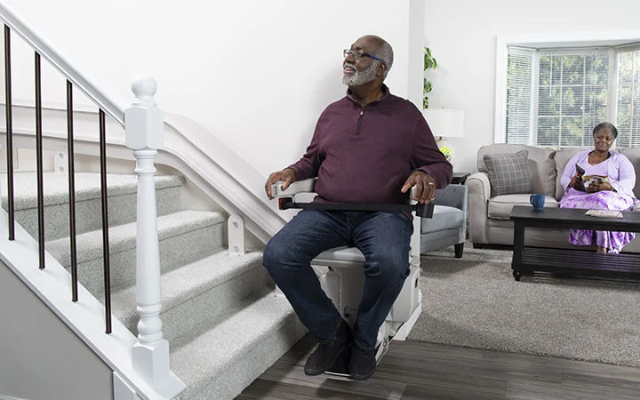 a man riding a curved stair lift with his wife sitting in the living room behind him watching