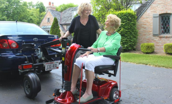 An elderly woman parking a scooter on a scooter lift mounted on the back of a blue car