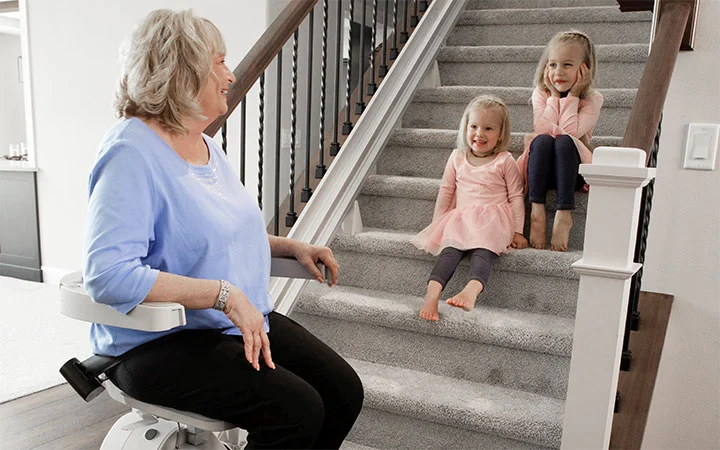 a woman riding an indoor stair lift with kids sitting on the steps watching
